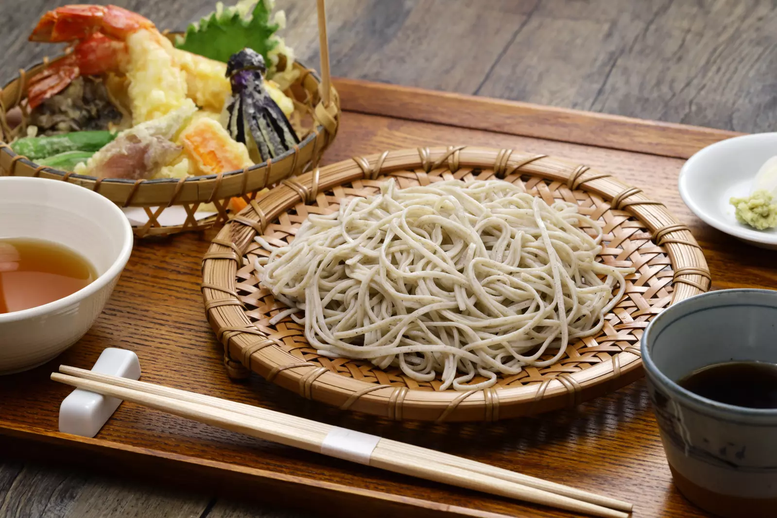 A serving of Japanese soba noodles on a bamboo tray with dipping sauce, tempura vegetables and shrimp, chopsticks, and wasabi on the side.