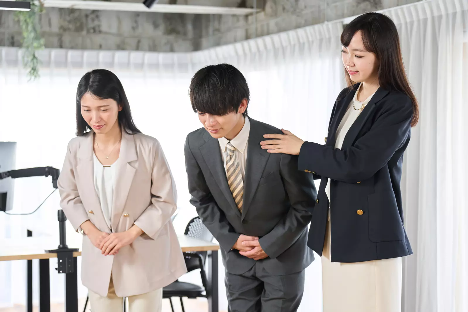 Three Japanese business people in formal attire, with two bowing while one places a hand on a colleague's shoulder, possibly during etiquette training for saying thank you.