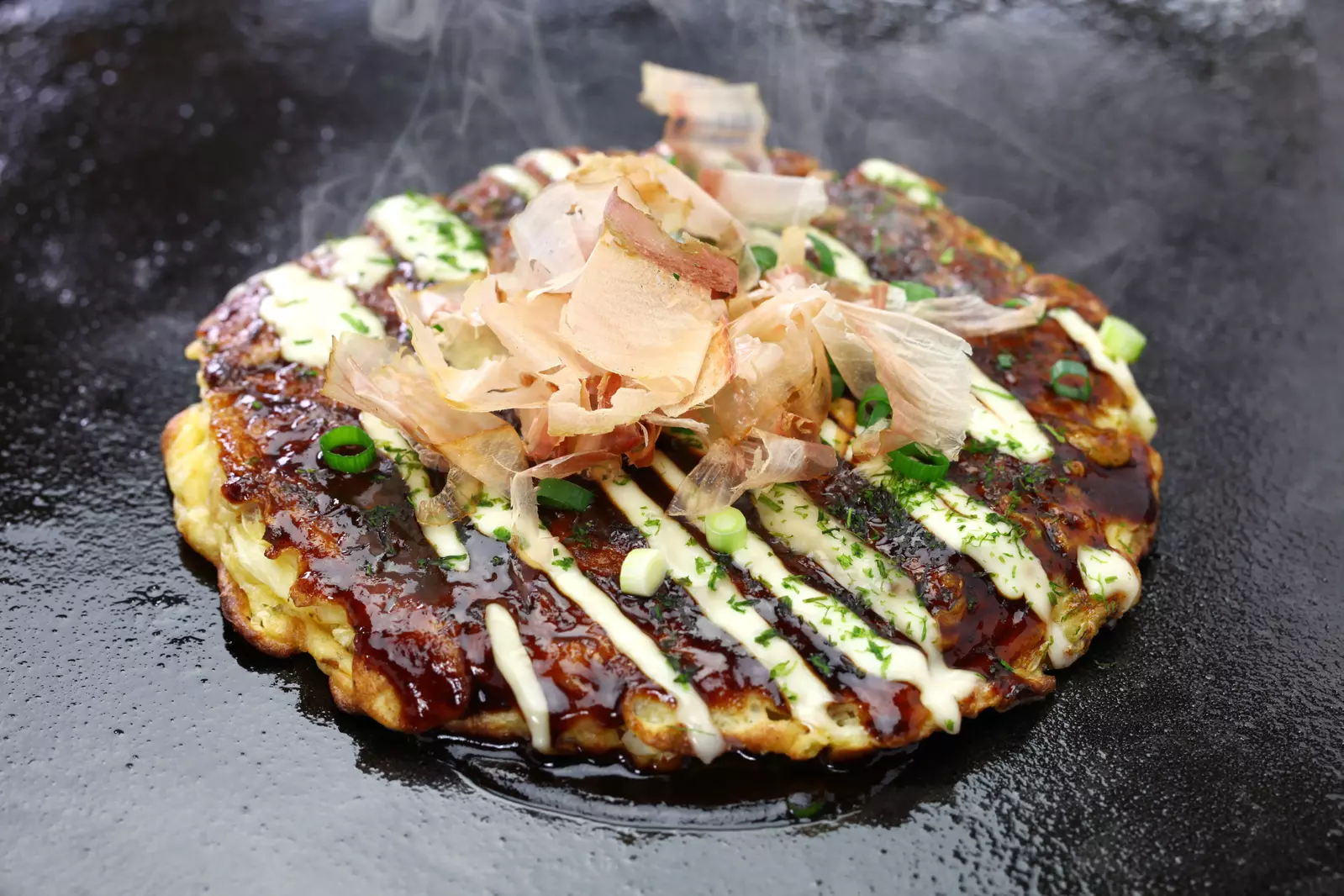 A freshly cooked Japanese okonomiyaki topped with bonito flakes, green onions, mayonnaise, and a savory sauce on a griddle.