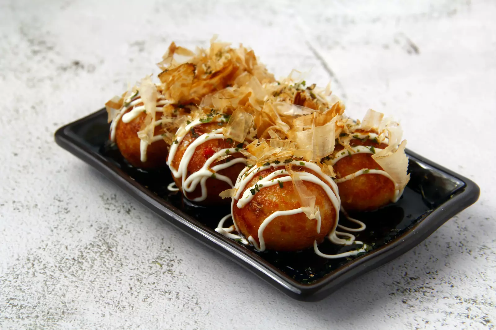 Japanese takoyaki, octopus-filled dough balls, topped with bonito flakes, mayonnaise, and sauce, served on a black plate.