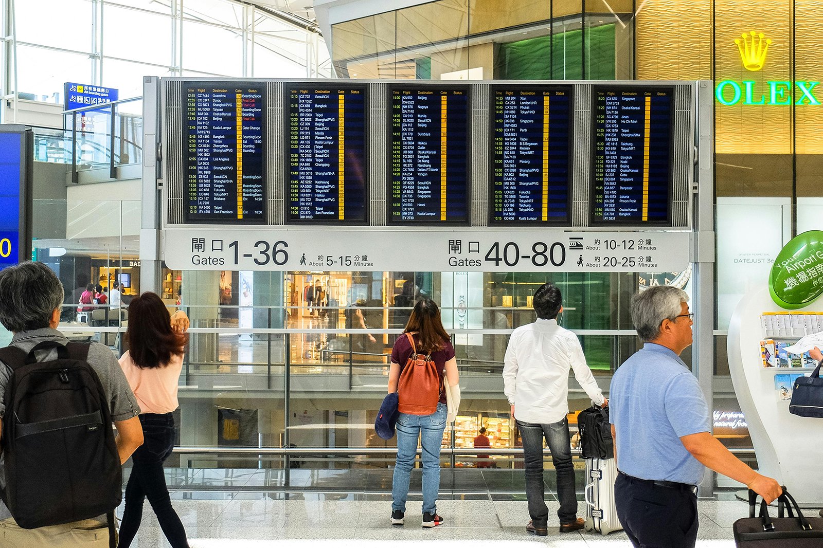 A screenshot showing several people standing in an airport lobby, looking up at the arrivals and departures board